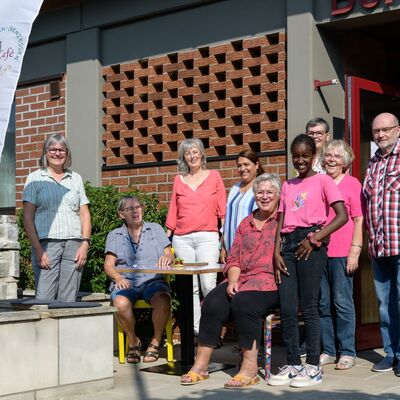 Mehrere Männer und Frauen unterschiedlichen Alters und ein Mädchen stehen und sitzen für ein Gruppenfoto auf der Terrasse eines Café, davor ein Aufsteller mit dem Namen EllCafé