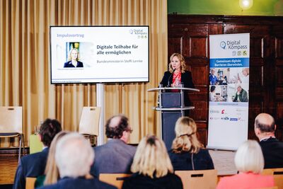 Ministerin hält Rede am Stehpult, Mensche sitzen im Saal davor und hören zu.