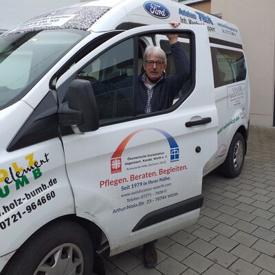 Grauhaariger Mann mit Brille und Schnäuzer schaut durch das geöffnete Seitenfahrerfenster eines mit bunten Aufklebern versehenen Transporter-Busses