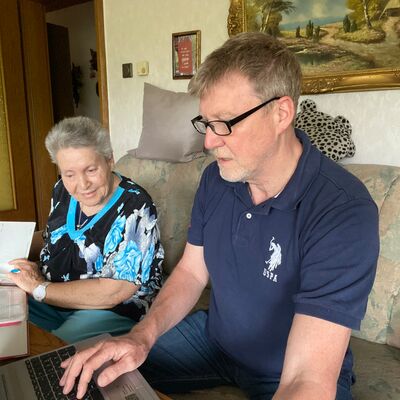 Mann mit Brille sitzt auf Sofo und tippt ins Notebook vor ihm, älter Frau sitzt neben ihm und schaut zu.