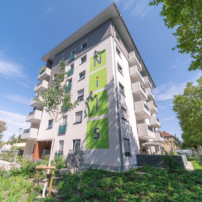 Sechsstöckiges Wohngebäude mit Balkonen auf der Wand ein großer Schriftzug NILS, rundum Grünanlagen mit grünen Pflanzen und Bäumen