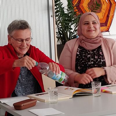 Bild mit zwei lachenden Frauen, eine Frau mit einem Buch, daneben eine Frau mit Migrationshintergurnd