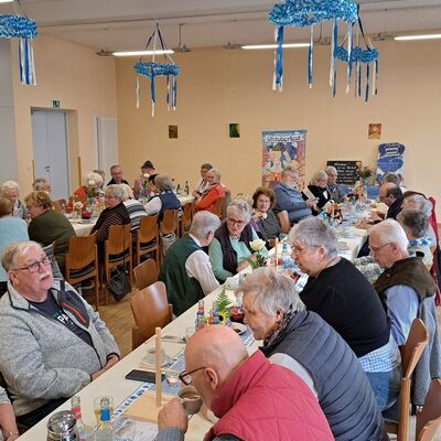 an langen Tischtafeln die bayerisch blau-weiß geschmückt sind sitzen Senioren beim Speisen und Getränken. In der Halle hängen einige blau-weiße Kränze von der Decke