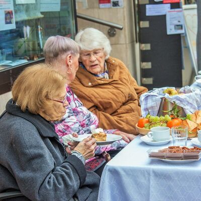 Drei Damen unterhalten sich bei Kaffee und Kuchen und Obst an einem Tisch im Außenbereich.