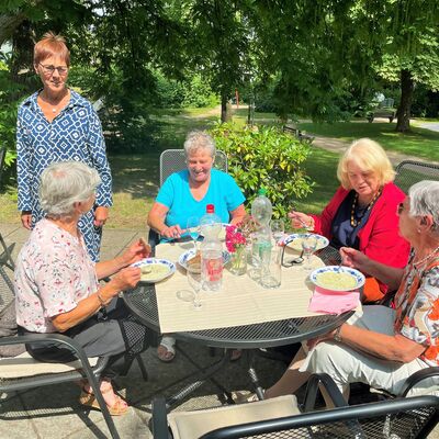 Vier Frauen sitzen am Tisch im Freien, unterhalten sich und essen Suppe, eine weitere Frau stüßt dazu