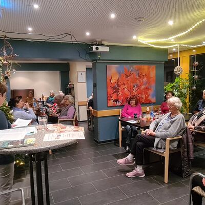 In einem weihnachtlich geschmückten Raum sitzen ältere Menschen und blicken auf eine vorlesende Frau an einem Stehtisch.