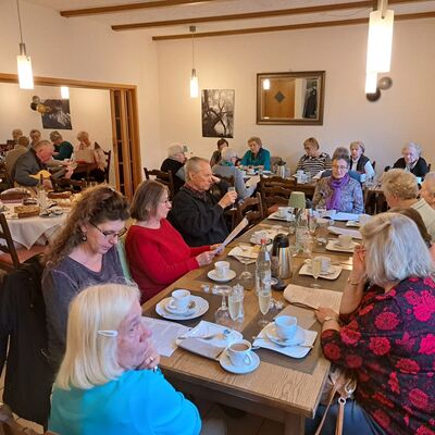 2 Gruppen mit überwiegend älteren Frauen sitzen über Eck an zwei gedeckten Kaffetischtafeln, im Hintergrund spielt ein alter Mann Gitarre, daneben ein Tisch mit diversen Kuchen und Torten