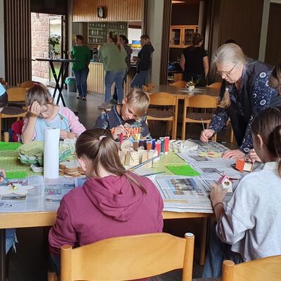 fünf Kinder und zwei Frauen sind an einem Gruppentisch mit Malarbeiten beschäftigt.