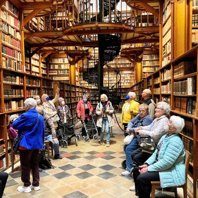 Frauen und Männer sitzen mit Mund-Nasenschutz-Masken in hohem Bibliotheksraum mt Wandregalen voller Bücher