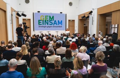 Saal mit ettlichen Menschen, die einer Rednerin zuhören, eine Kamera filmt diese.