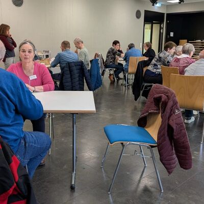 Männer und Frauen sitzen in einem Raum mit mehreren Tischen, unterhalten sich, essen oder spielen miteinander.