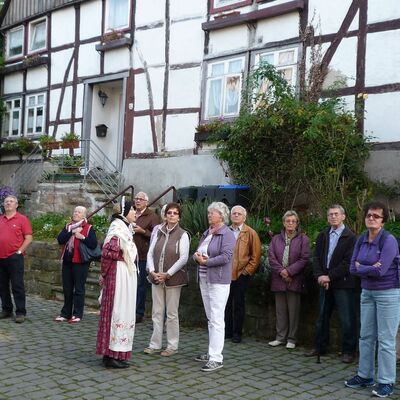 Bild mit einer Seniorengruppe, die vor einem Fachwerkhaussteht, eine Frau in Tracht erklärt etwas.