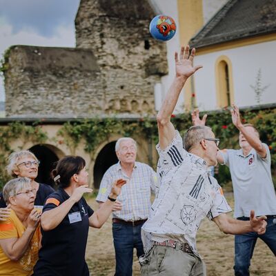4 Frauen und 3 Männer auf einem Sandplatz vor einer Klosterkirche bei einem Ballspiel. Ball hoch oben in der Luft, eine Frau und 2 Männer haben die Arme nach oben gestreckt