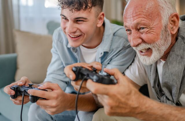 Bild mit jungem und alten Mann die auf einem Sofa sitzen und lachend Spielekonsolen in den Händen halten
