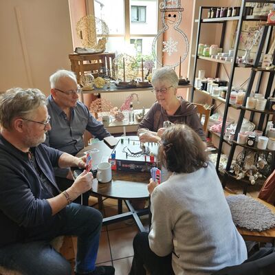 Bild mit je zwei älteren Männern und Frauen an einem Tisch im Ladeninterieur mit Regealen. Sie halten Karten in der Hand auf der Tischmitte ein Spielbrett sowie Tassen.