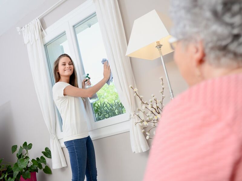Junge Helferin beim Fensterputzen im Wohnzimmer einer älteren Dame