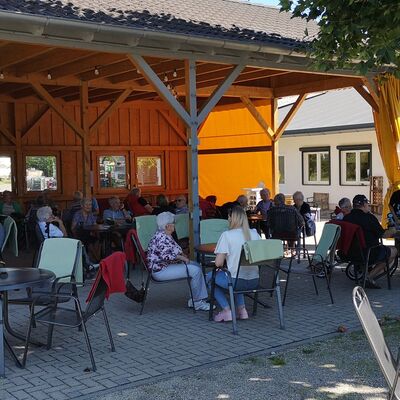Vorwiegend ältere Männer und Frauen sitzen in Gruppen auf einer überdachten Terasse mit Außenmöbeln und zusammengeklappten Sonnenschirmen