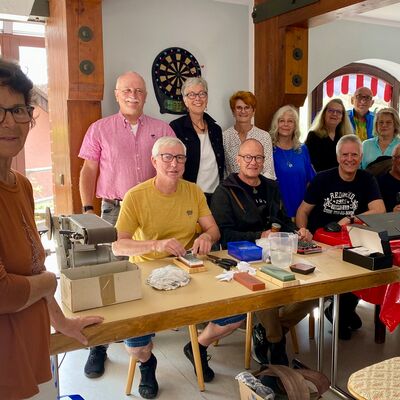 Gruppenbild mit zwölf Frauen und Männern, die an einem Tisch mit Nähmaterial und anderen Reparatur-Utensilien sitzen.