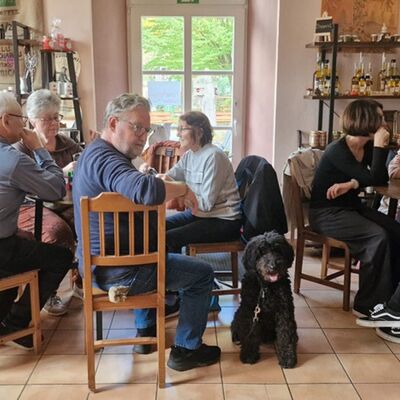 Bild aus einem Ladengeschäft in dem zwei Tische stehen um die herum Männer und Frauen beim Spiel sitzen, ein Hund im Vordergrund
