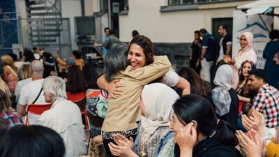 Bild mit Menschen unterschiedlichen Alters und Herkunft sitzen in einem Hof zusammen, zwei Frauen umarmen sich.