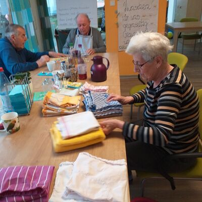 Eine alte Dame sitzt am Tisch und faltet Wäsche, im Hintergrund sitzen 2 alte Männer bei Tätigkeiten
