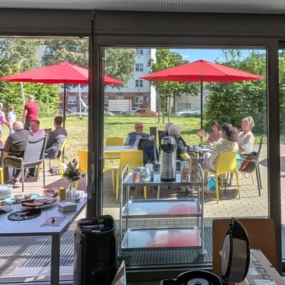Blick von einem Raum mit einem gedeckten Tisch durch ein Fenster auf eine Terasse im Grünen. Auf gelben Stühlen an Tischen unter roten Sonnenschirmen sithzen Menschen unterschiedlichen Alters