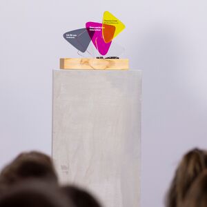 Foto mit einer Stele auf der ein Preis-Pokal steht, davor sitzen Menschen.