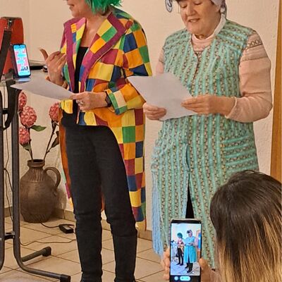 Frau im Anschnitt hält Smartfon hoch und nimmt Bild auf von den beiden Frauen, die verkleidet auf der Bühne stehen, links Frau mit grüner Perücke und bunt-kariertem Frack, recht Frau mit Kittel und Kopftuch, beide halten Zettel in der Hand mit einem Vortragstext