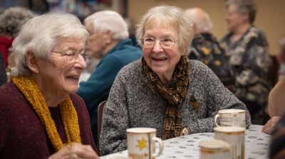 Zwei älter Frauen sitzen lachend mit Kaffeetassen an einem Tisch, im Hintergrund ein Tisch mit weiteren Seniorinnen und Senioren.