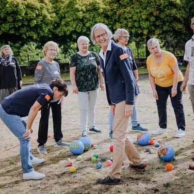 8 Frauen und 1 Mann auf einem Sandplatz, im Sand zwischen ihnen liegen bunte Bälle unterschiedlicher Größe.