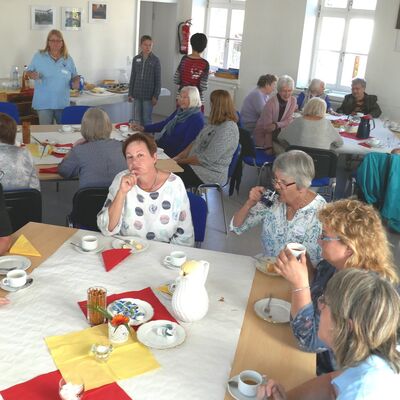 Männer und Frauen unterschiedlichen Alters sitzen an gedeckten Cafétischen und unterhalte sich, im Hintergrund ein Cafébuffet.