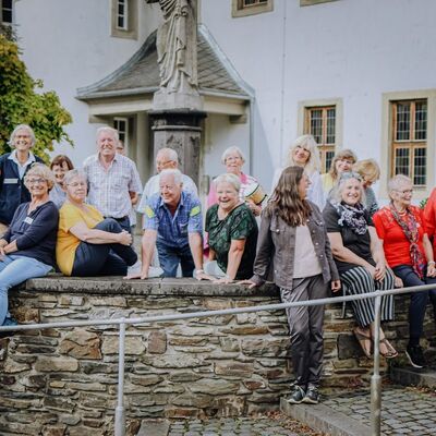 Gruppenbild mit Frauen und Männern unterschiedlichen Alters, die vor einem Klostergebäude hinter und auf einer Bruchsteinmauer stehen bzw. sitzen.