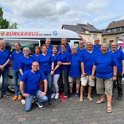 Gruppenfoto mit 16 Männern und Frauen die in dunkelbauen T-Shirts vor dem Kleinbus stehen.