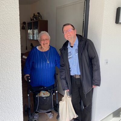 Mann steht mit Stoffbeutel an einer Haustür, die von einer lachenden älteren Frau am Rollator geöffnet wurde.