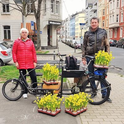 Frau und Mann mit stehen am Lastenrad, das mit Paletten gelb blühender Osterglocken beladen ist.