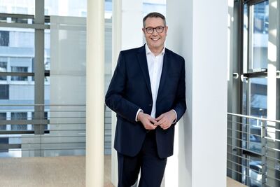 Minister Alexander Scheitzer mit Anzug und weißem Hemd, lachend an Säule gelehnt