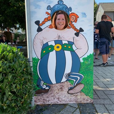Bild mit Frau, die durch ein ausgespartes Gesicht eines auf einer Holztafel aufgemalten Comicfigur Obelix in Lebensgröße blickt.