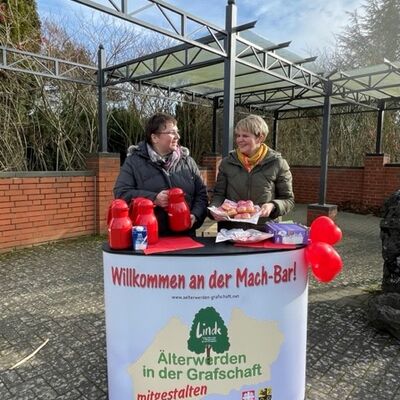 Zwei Frauen mit Jacken hinter einer Theke im Freien auf der rote Thermaoskannen stehen. Vorna der Theke das Projektlogo sowie der Hinweis "Willkommen an der Mach-Bar!"