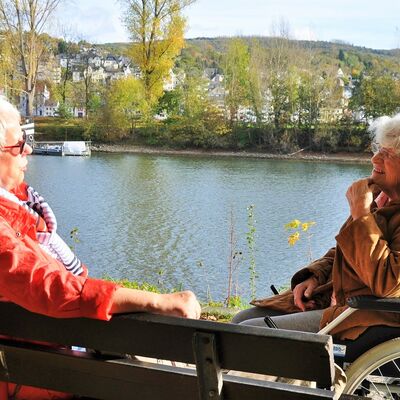 Dame auf Parkbank und Dame im Rollstuhl sitzen sich unterhaltend am Rhein.