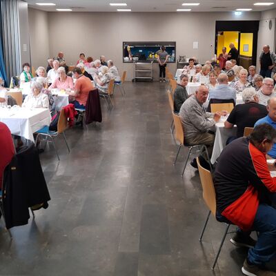 langezogener Raum mit zehn gedeckten Tischen, an denen rund 50 bis 60 Seniorinnen und Senioren sitzen, im Hintergrund eine Durchreiche zu einer Küche