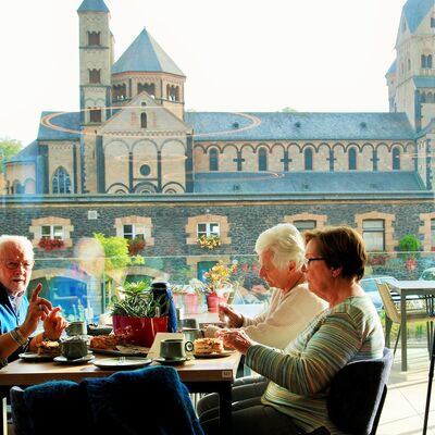 Männer und Frauen sitzen am Cafetisch vor einem großen Glasfenster, im Hintergrund die Klosterkirche Maria Laach