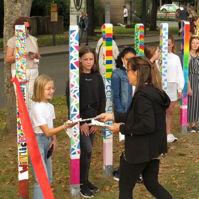 Bild von einer Straße mit Bäumen an denen bunt gestaltete Pfähle aufgereiht sind, dazwischen stehen Menschen verschiedenen Alters