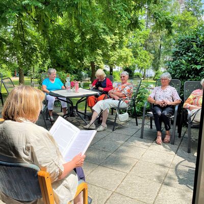 Ältere Frauen sitzen auf Terassenstühlen im Grünen und hören einer Vorleserin zu