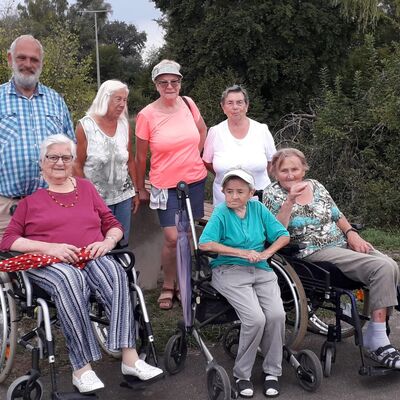 Gruppenfoto im Freien vor Büschen und Bäumen: 3 ältere Damen im Rollstuhl im Vordergund, ein älterer Mann und 3 ältere Frauen im Hintergrund w