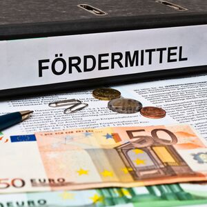 Bild mit einem Aktenordner auf dem Fördermittel steht. Davor liegen Formulare, ein Kugelschreiber sowie Papiergeld und Münzen