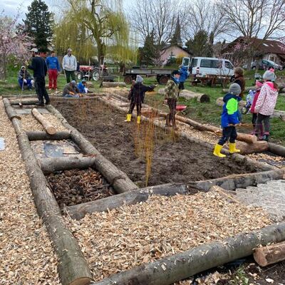 Kinder, Männer und Frauen warm angezogen im Frühling. Auf dem Boden liegen Baumstämme die einen Parcours abstecken, verschiedene Räume sind mit Holzspänen, Steinen, Erde und anderen Materialien gefüllt