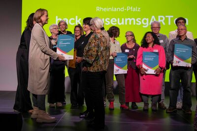 Bild mit der Ministerin die Urkunde an eine Frau übergibt. Auf der Bühne viele weitere Menschen mit Urkunden