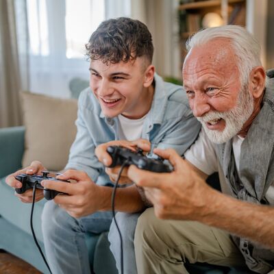Bild mit jungem und alten Mann die auf einem Sofa sitzen und lachend Spielekonsolen in den Händen halten