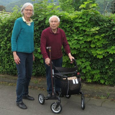 Alter Mann am Rollator, an dem ein Stock befestigt ist auf der Straße, neben ihm eine ältere Frau.