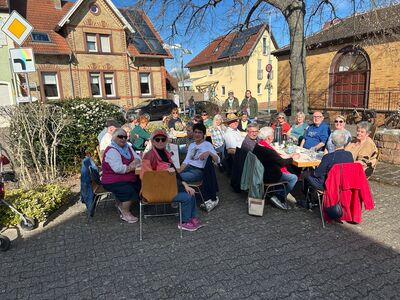 Bild mit einer Gruppe von Menschen die auf Stühlen an zwei gedeckten Tischen bei blauem Himmel im Freien in einem Ort sitzen.